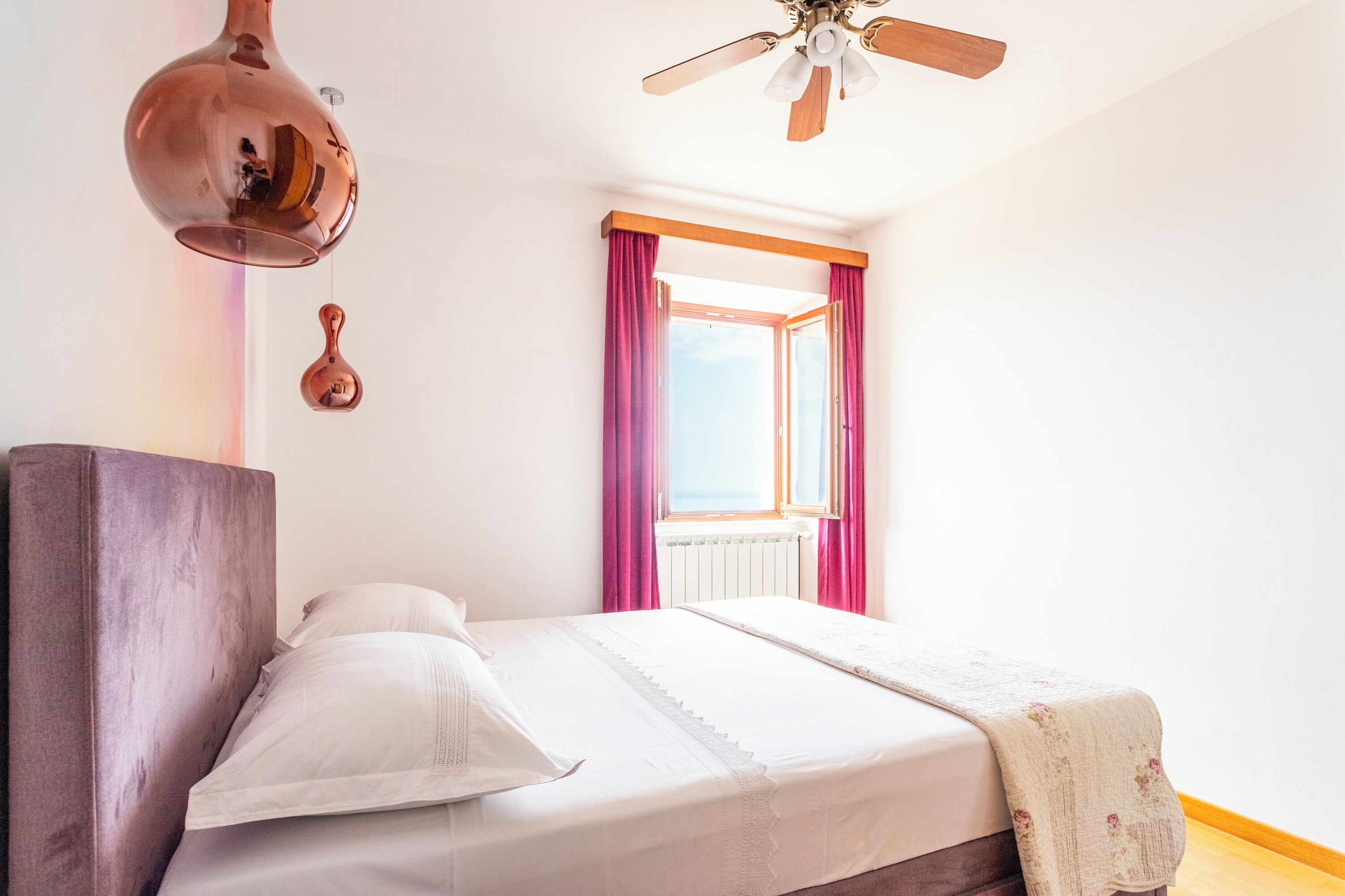 Quarto com cama, ventilador de teto, janela com cortinas e radiador.