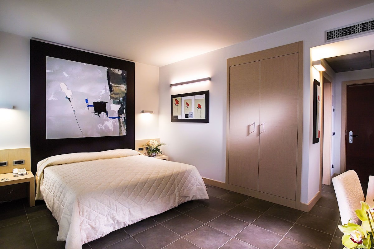 Ein Hotelzimmer mit einem großen Bett, einem Kleiderschrank, einem Schreibtisch mit Telefon und einem an der Wand hängenden Kunstwerk.