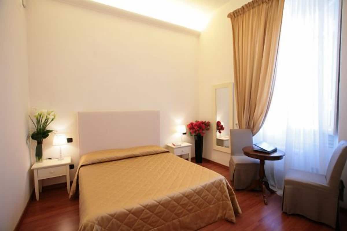 A hotel room with a large bed, a bedside table with a lamp, a chair, a small table, and a mirror.