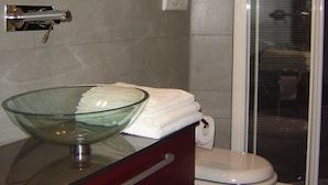Un baño con lavamanos de vidrio, inodoro blanco y cabina de ducha con paredes de vidrio.