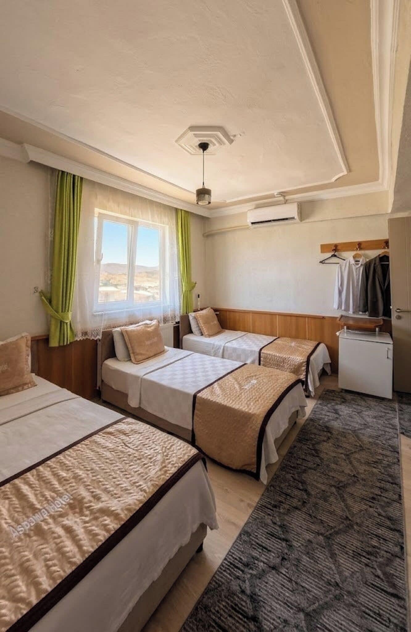 Habitació d'hotel amb dos llits, un escriptori petit, un frigorífic i una finestra amb cortines.