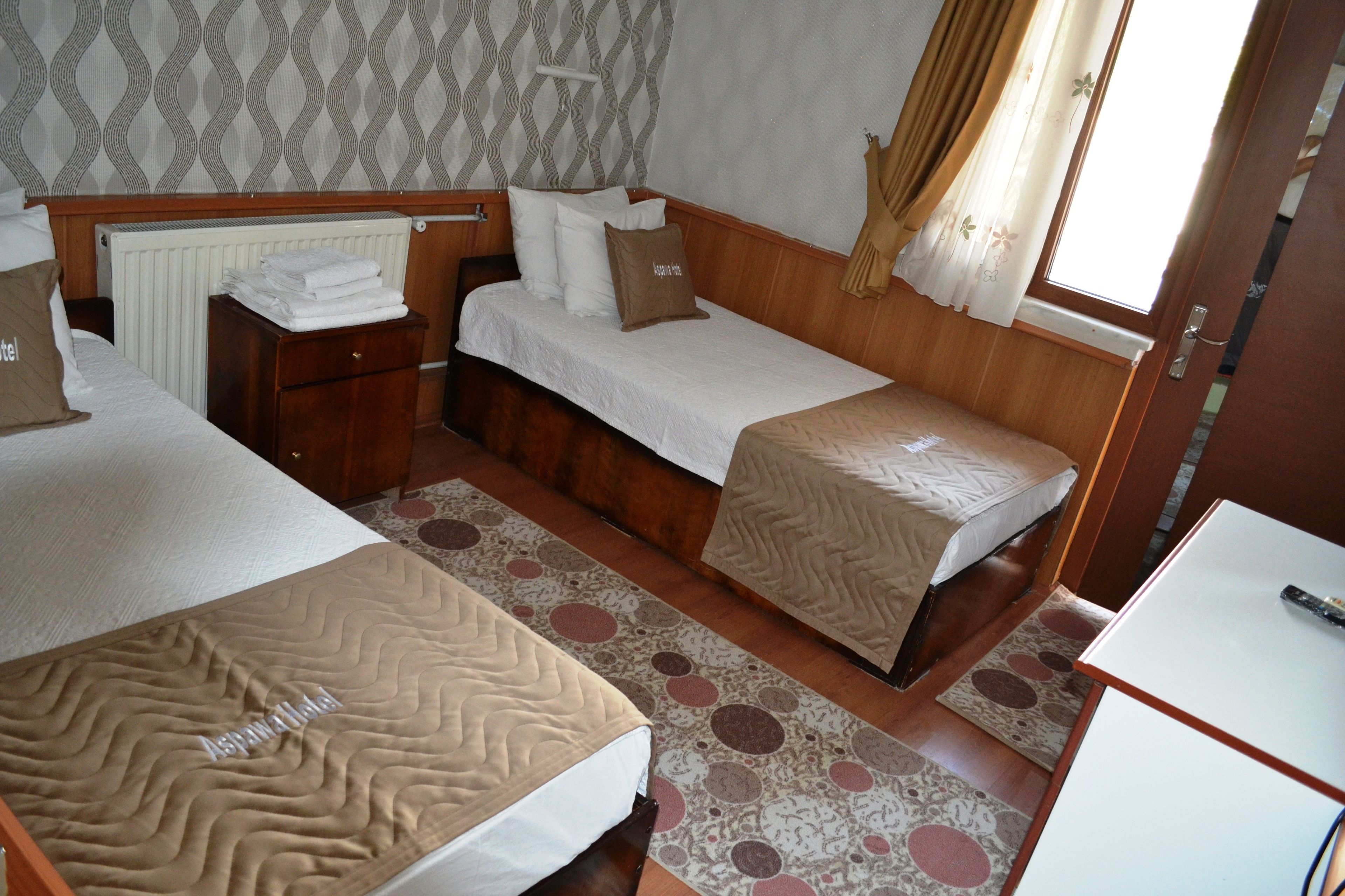 A hotel room with two beds, a wooden headboard, a radiator, a window with curtains, and a small desk.