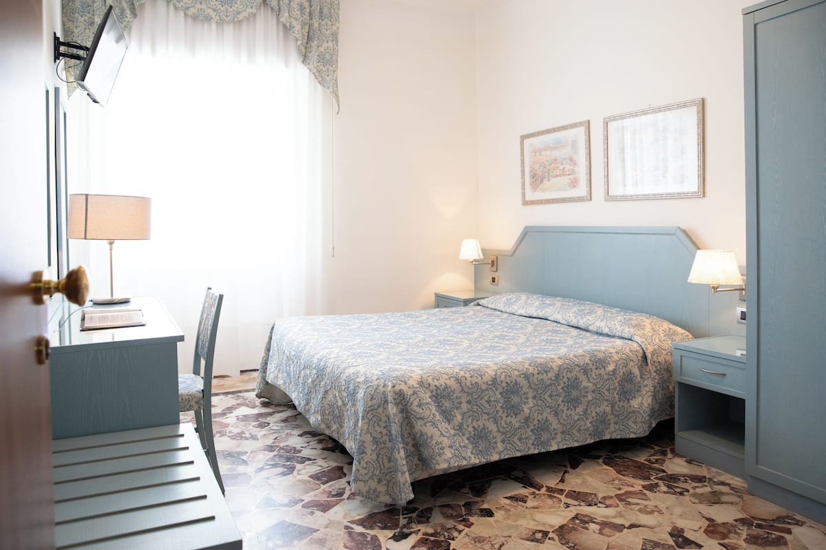 A bedroom with a bed, a desk, and a chair. There are two bedside tables with lamps. The room has a patterned floor and a window with curtains.