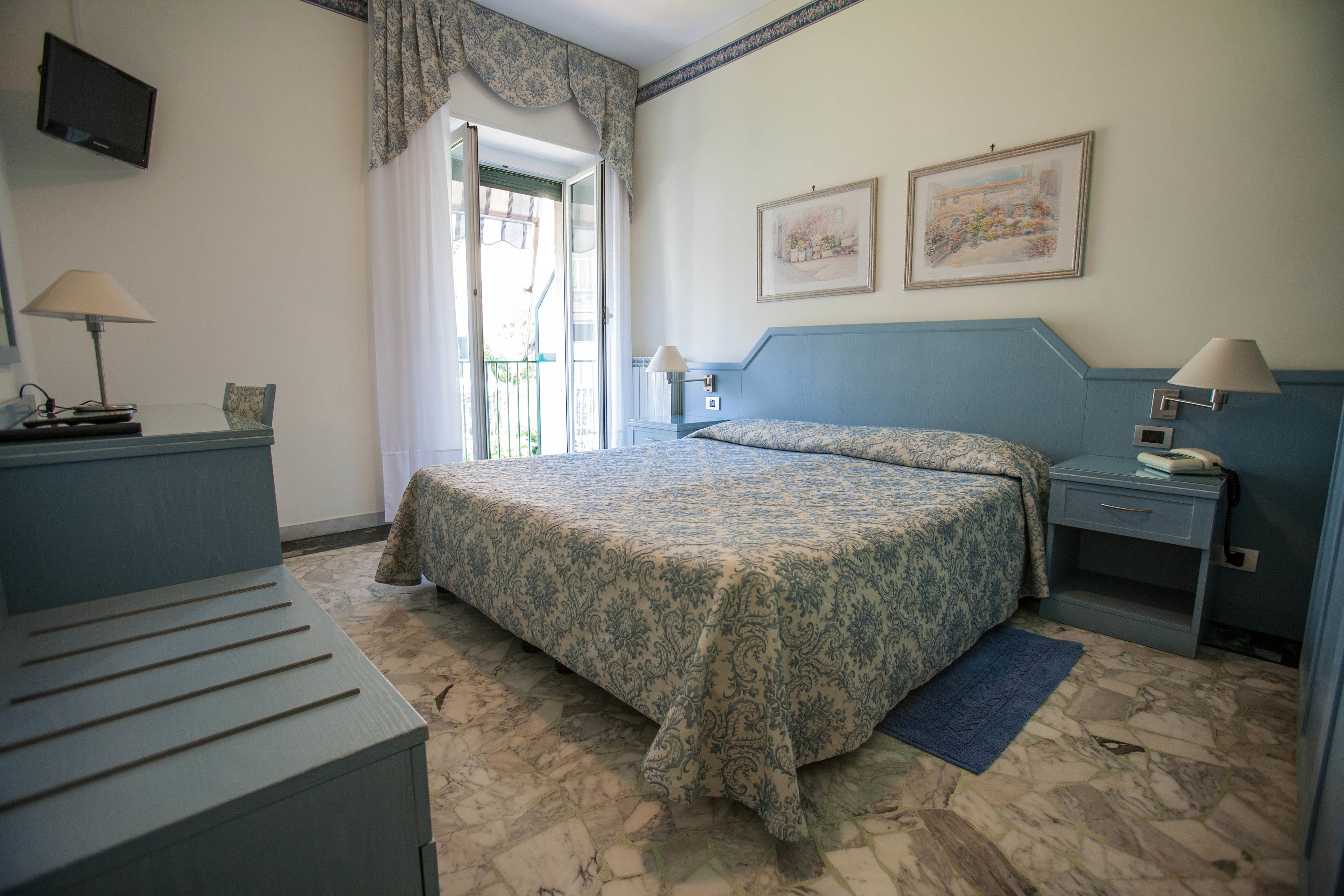 Un dormitorio con cama, escritorio, silla, mesita de noche, lámpara, televisor y ventana con cortinas.
