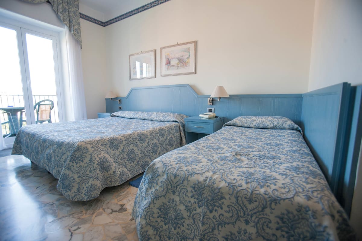 A hotel room with two beds, a nightstand, a lamp, and a window with a view.