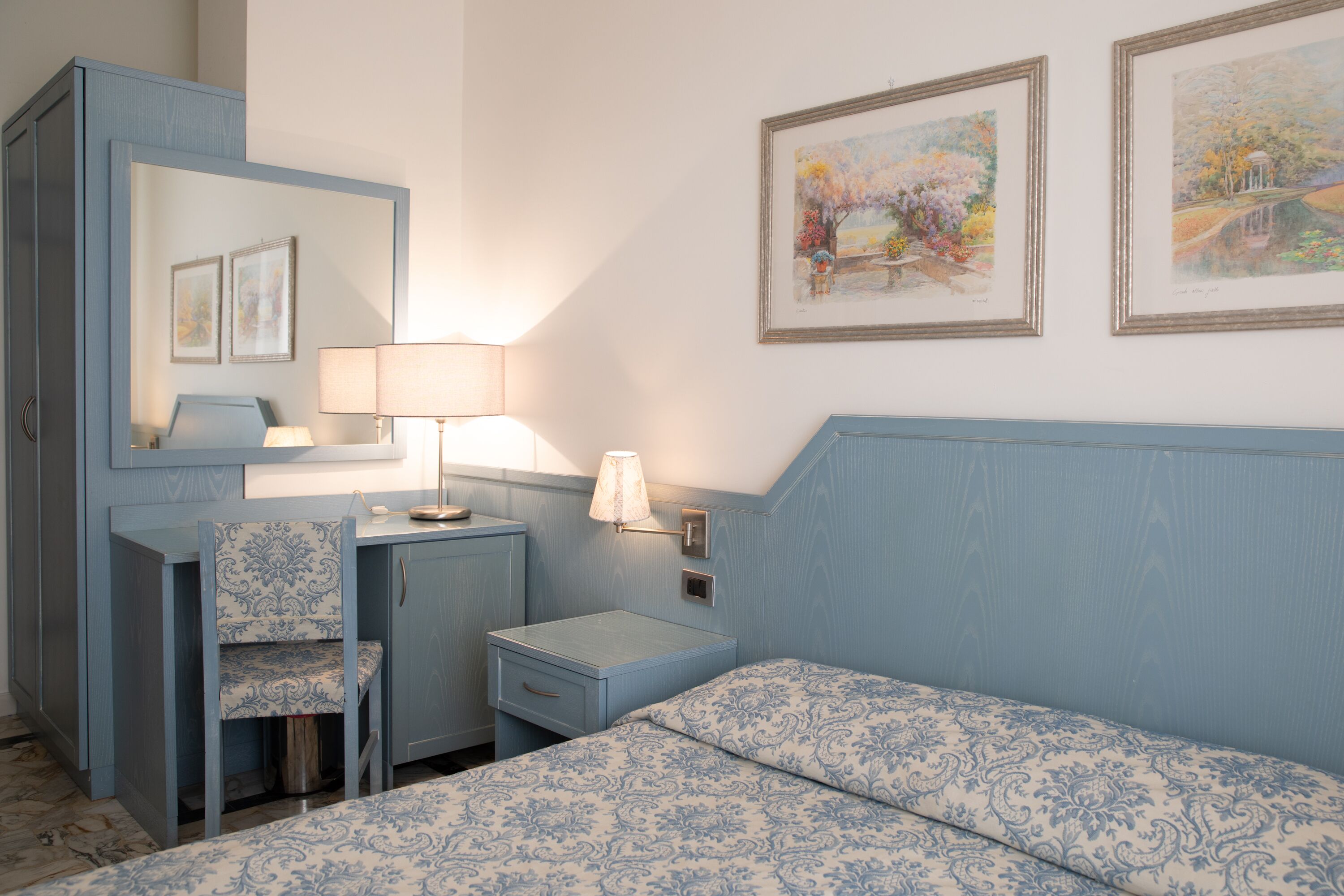 A bedroom with a bed, a desk, a chair, and a nightstand. There are two framed pictures on the wall.