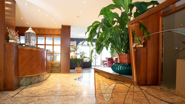 A lobby with a marble floor, wooden reception desk, large potted plants, and a view of the outdoors through large windows. - Hotel Ariston (Imperia)