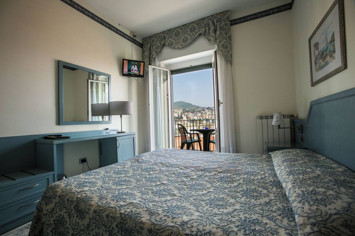 A bedroom with a bed, a desk, a chair, a TV, and a balcony with a view of the city.