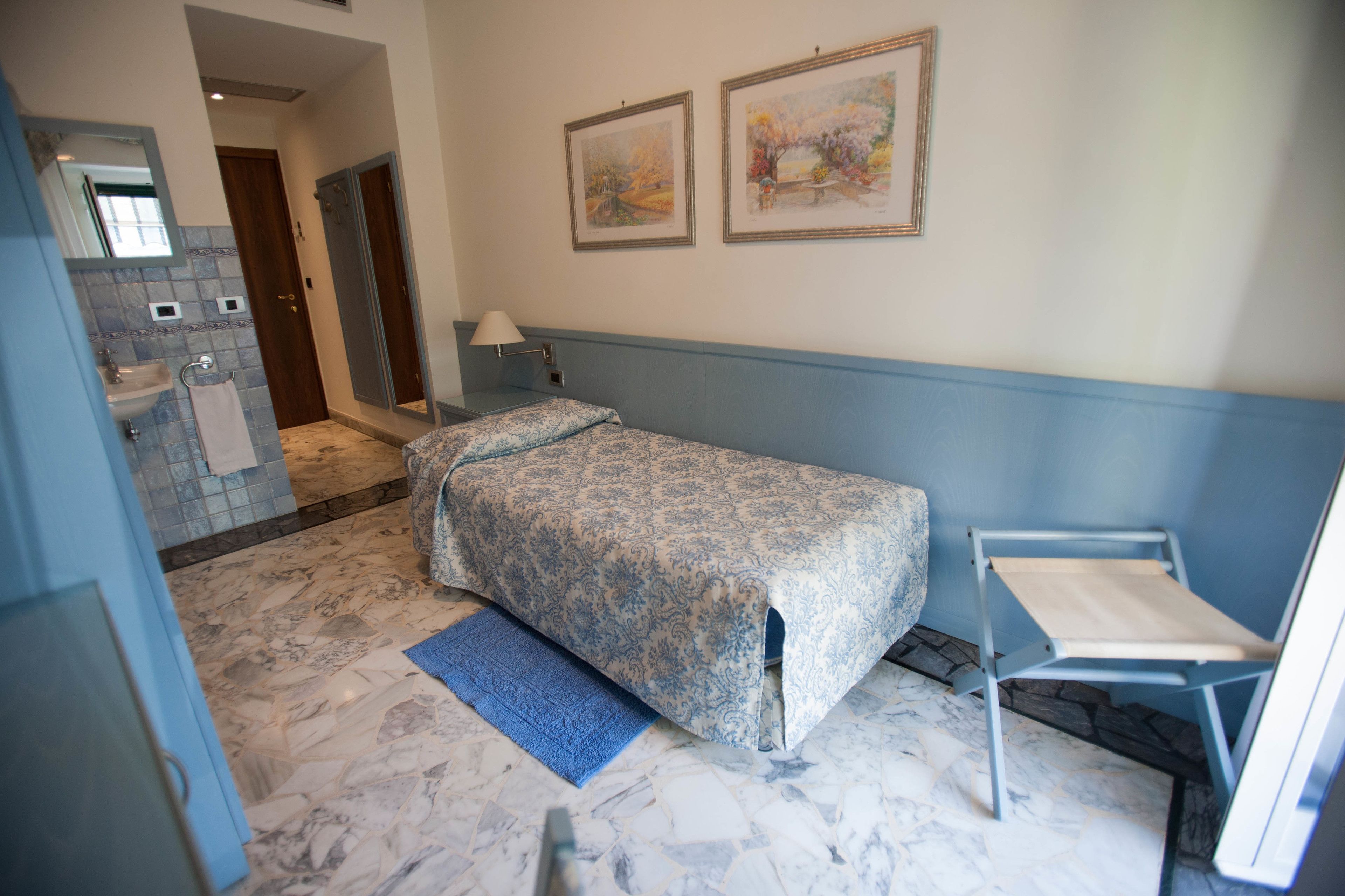Une chambre à coucher avec un lit, une chaise, une table de chevet, une lampe, et une salle de bain visible au fond.