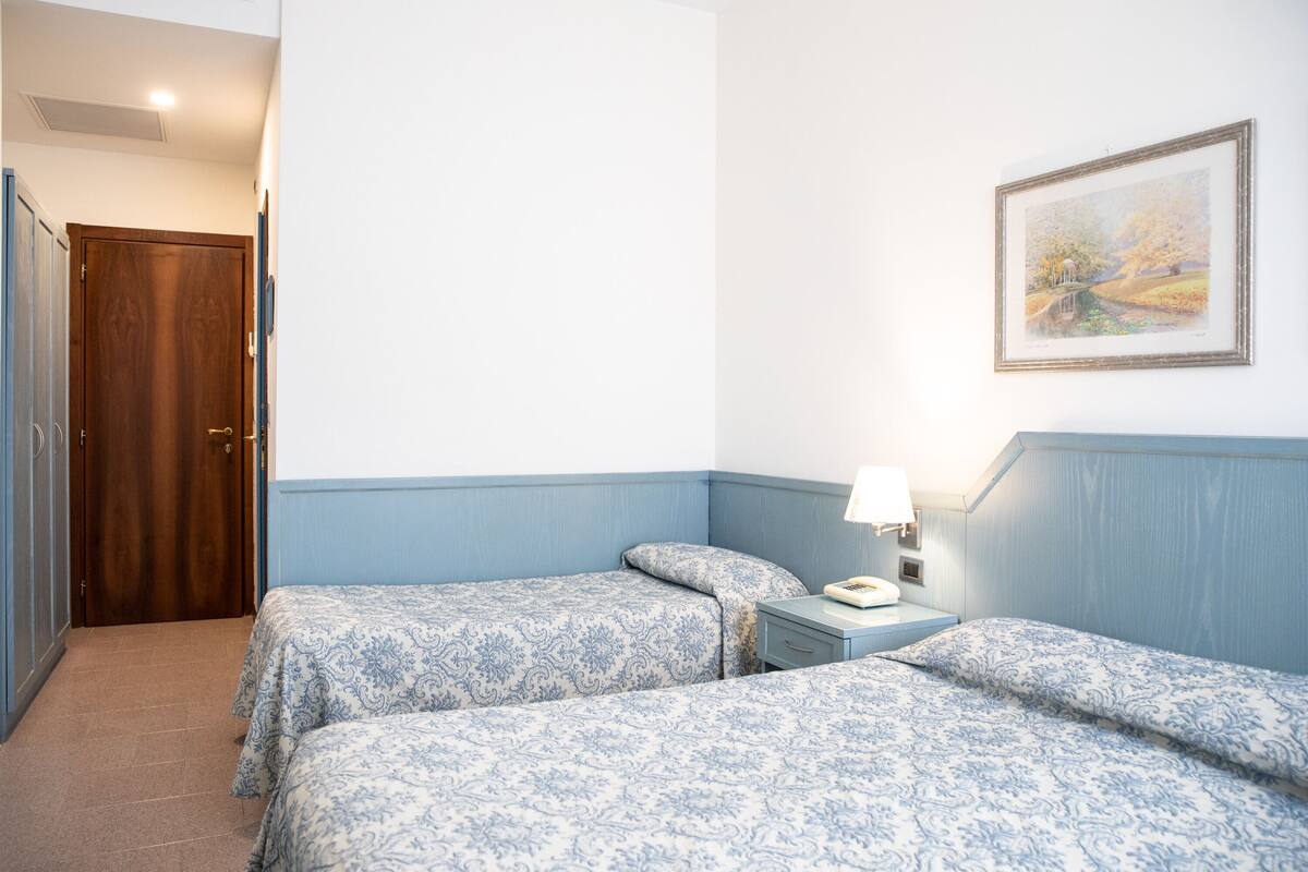 A hotel room with two beds, a wooden door, a bedside table with a lamp, and a framed painting on the wall.