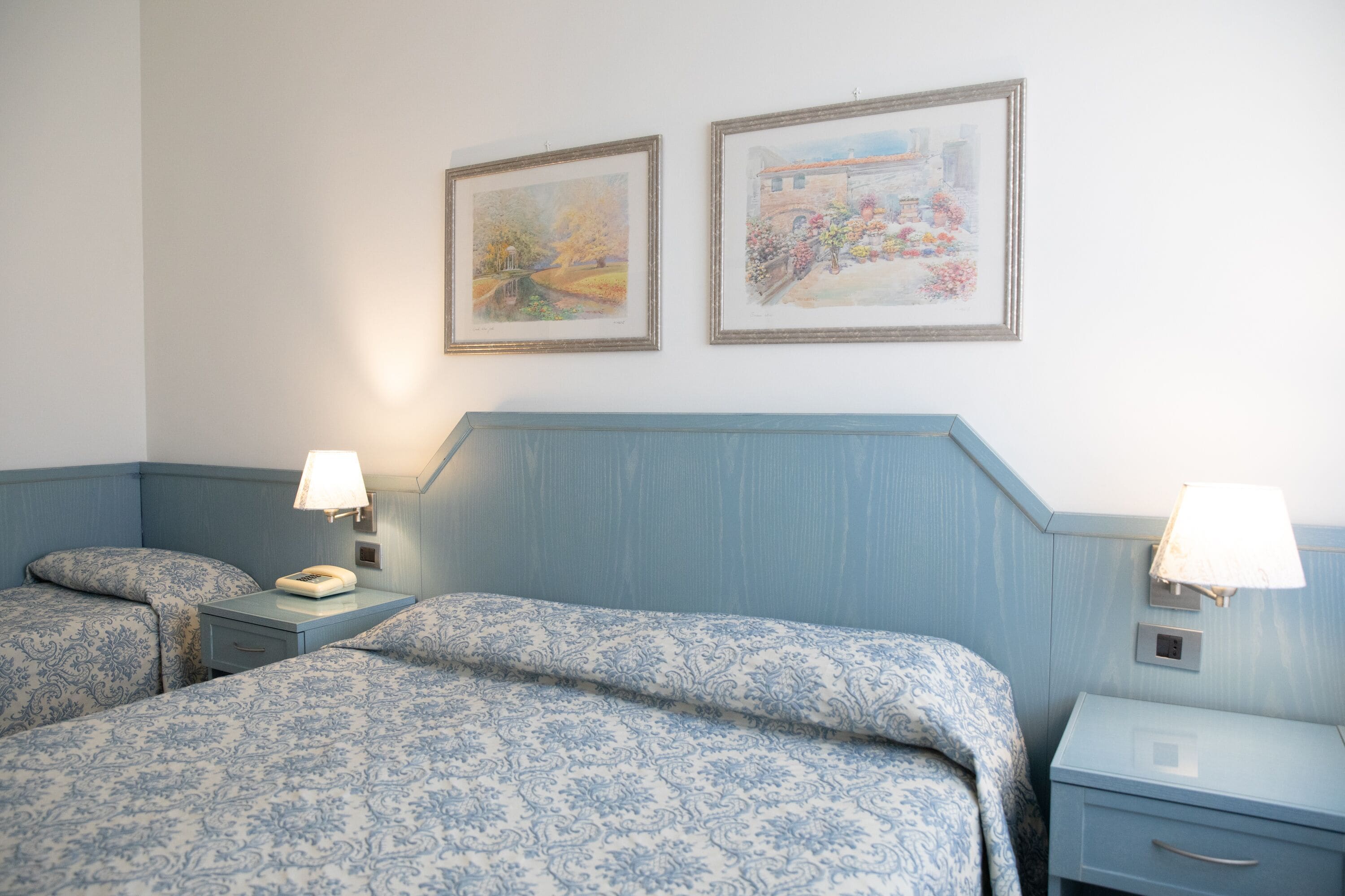 A hotel room with two beds, each with a patterned bedspread, a bedside table with a lamp, and two framed pictures on the wall.