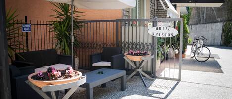 An outdoor seating area with wicker furniture, a potted plant, and a sign for 'Hotel Ariston'.