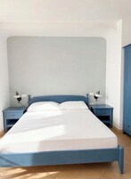 Un letto con struttura blu e biancheria bianca, affiancato da due comodini con lampade, in una stanza con una parete spoglia e un armadietto blu.