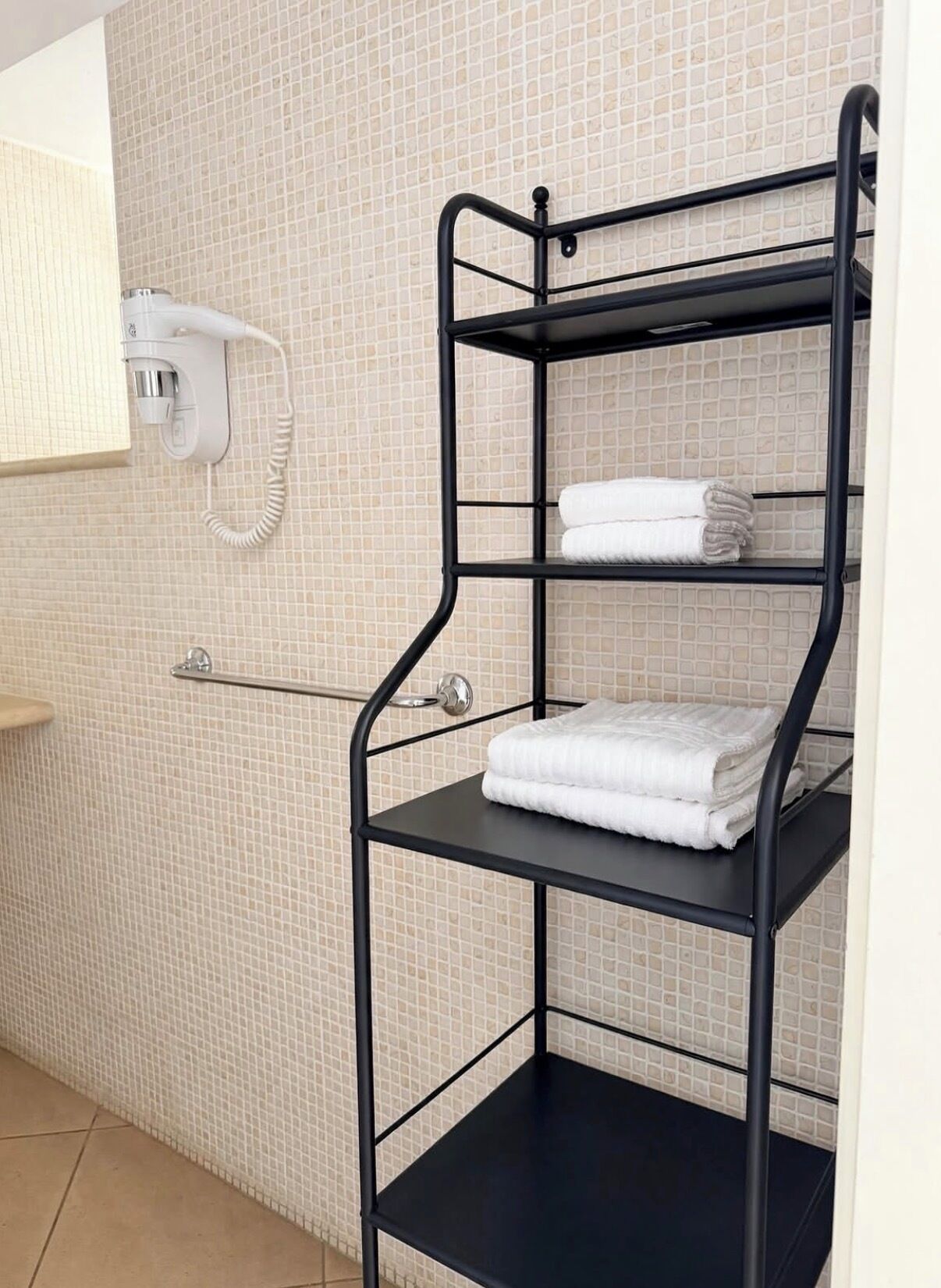 Una scaffalatura in metallo nero con asciugamani, un termosifone elettrico bianco e un telefono fissato alla parete in un bagno rivestito di piastrelle.