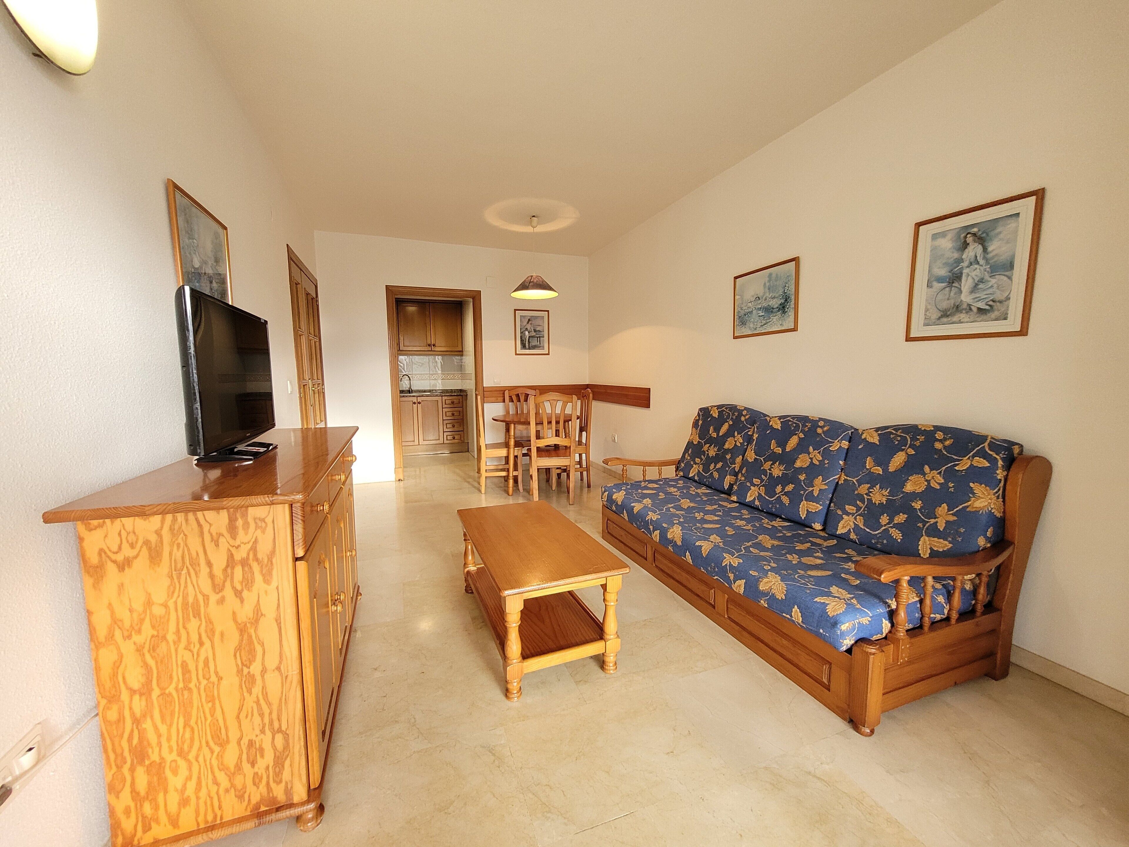 Olohuoneessa on puinen sohva, puinen TV-taso, ruokapöytä tuoleineen ja taustalla näkyy keittiö.