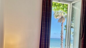 A hotel room with a bed, a lamp, a window with curtains, and a view of the sea and palm tree.