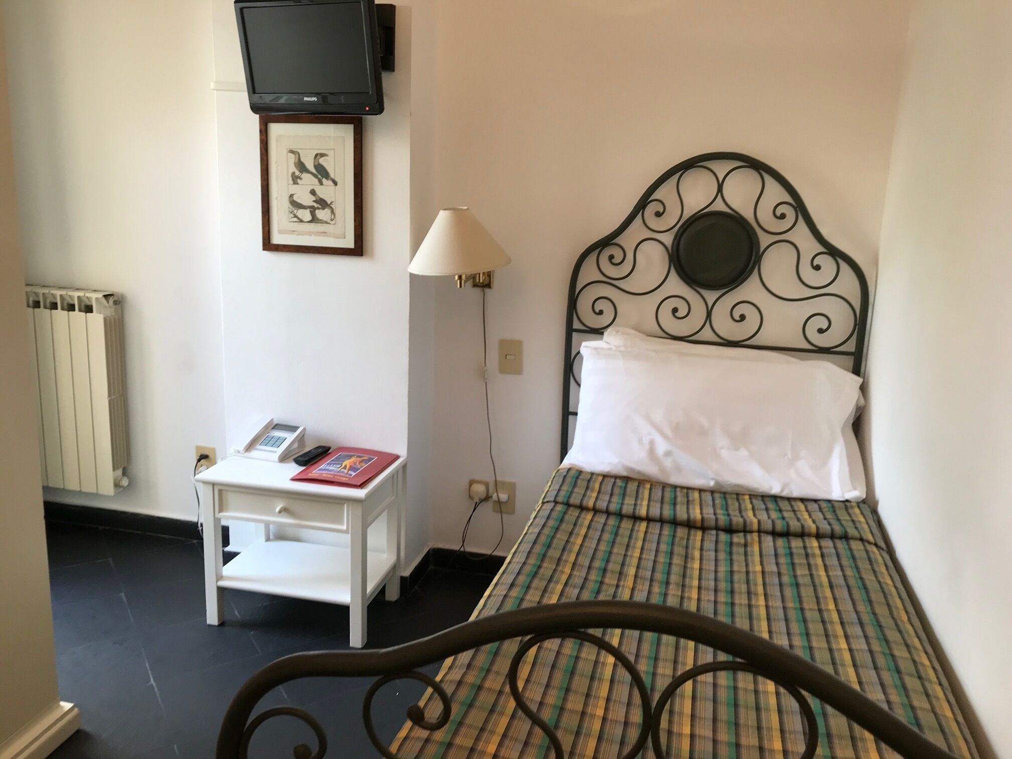 A small, neatly arranged bedroom with a single bed, a bedside table, a lamp, and a framed picture on the wall.