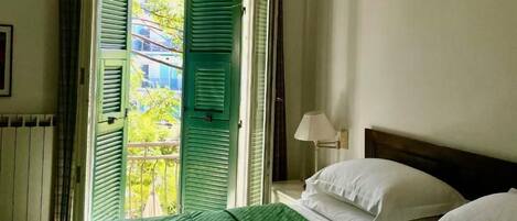 A room with a bed, a nightstand, a lamp, and a view through open shutters.