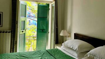 Una habitación de hotel con cama, mesita de noche con lámpara y ventana con contraventanas verdes.