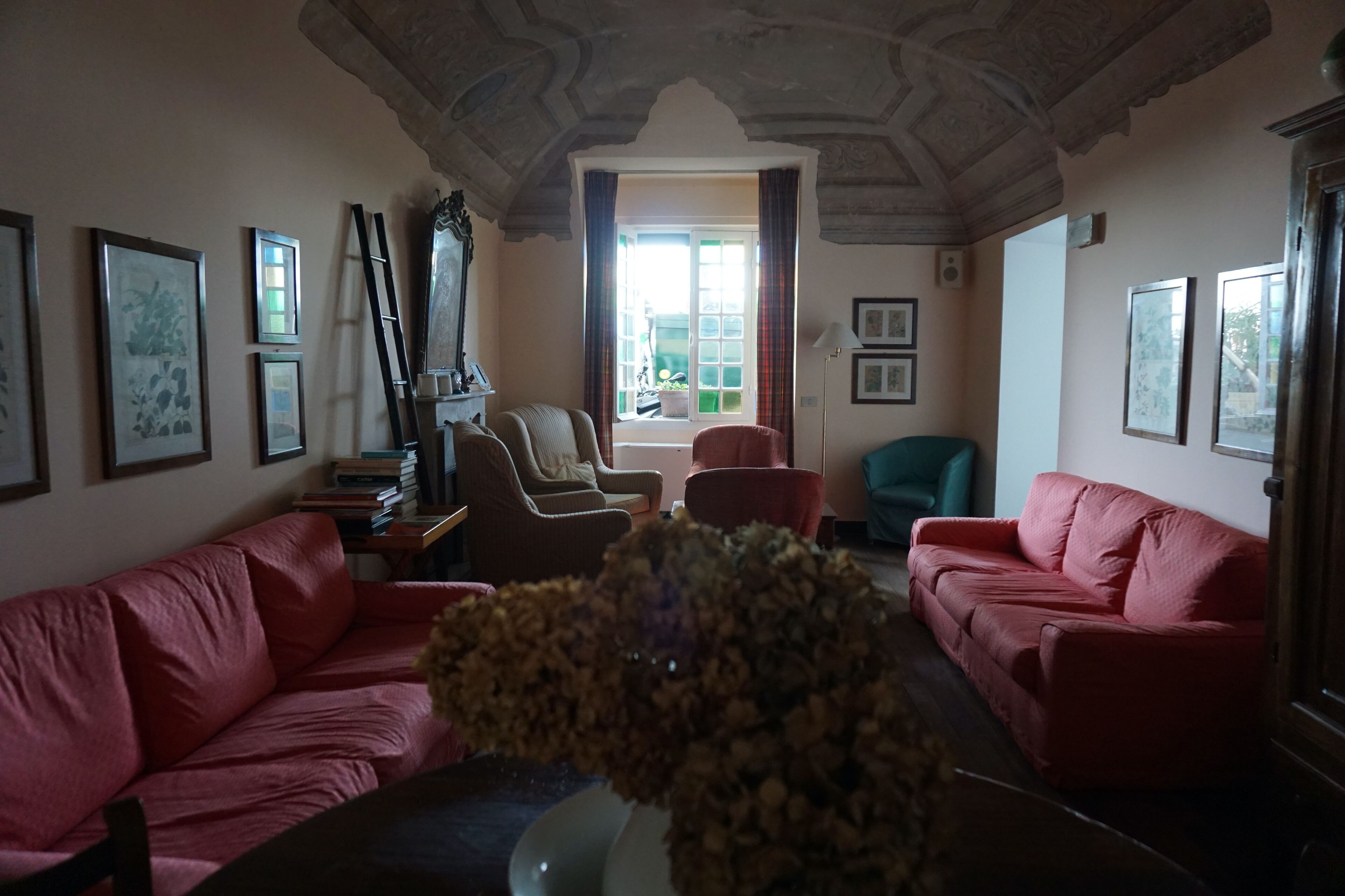 A living room with two red sofas, a coffee table, and a vase of flowers. There are framed pictures on the walls and a window with curtains.