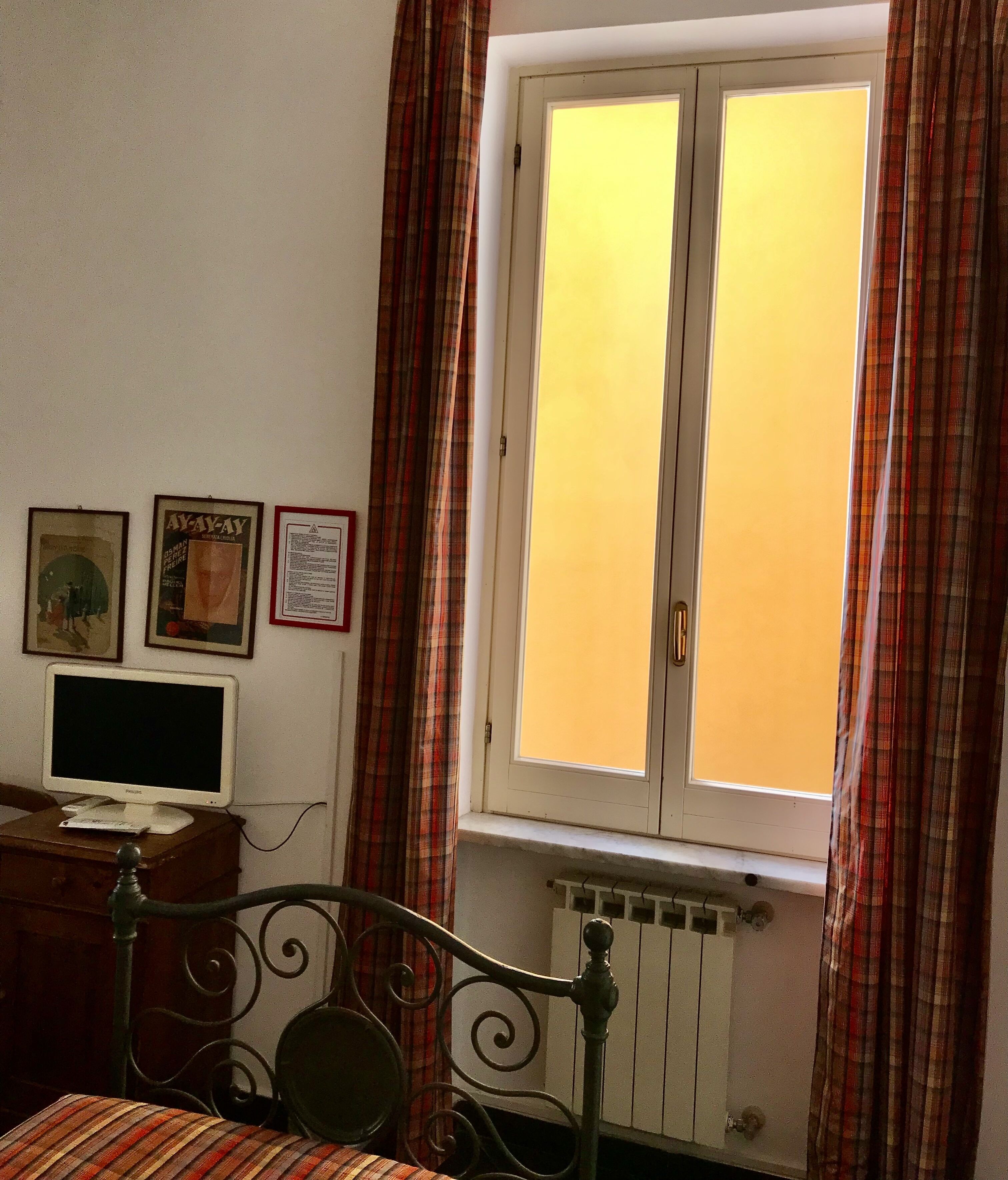 Un dormitorio con cama, mesita de noche, monitor y ventana con cortinas.