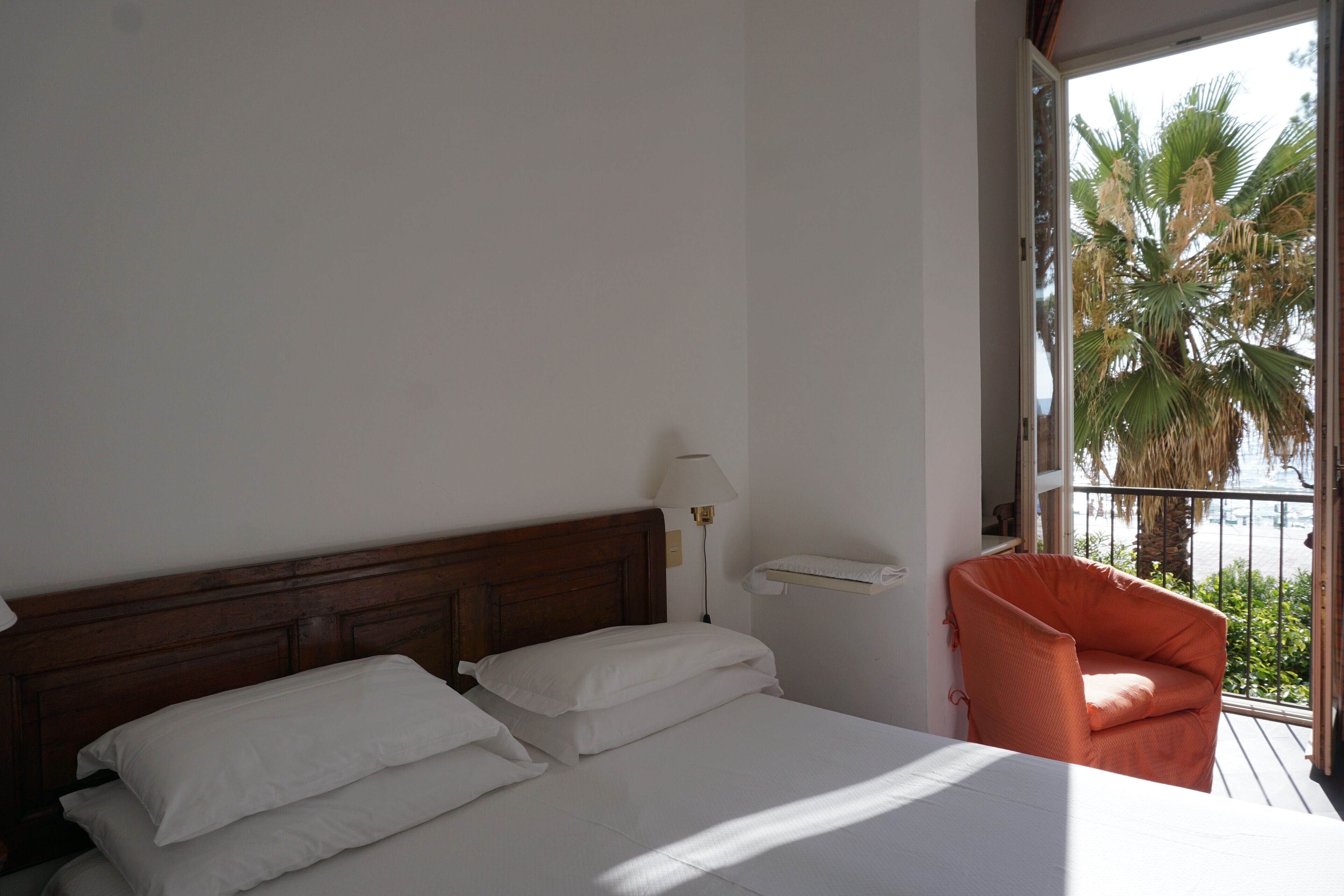 Un dormitorio con cama, mesita de noche, lámpara y silla junto a una ventana con vista a palmeras.
