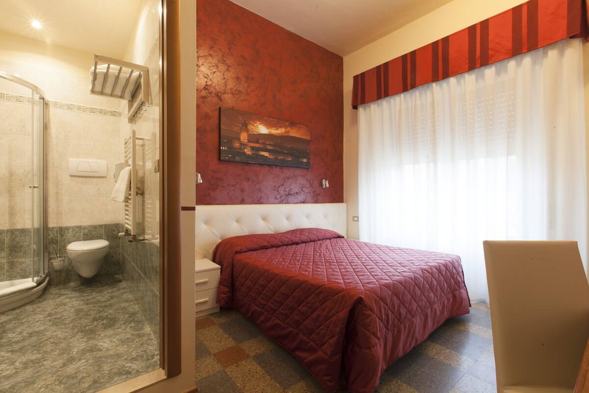 A hotel room with a glass shower enclosure, a bed with a red quilt, a white bedside table, and a chair.