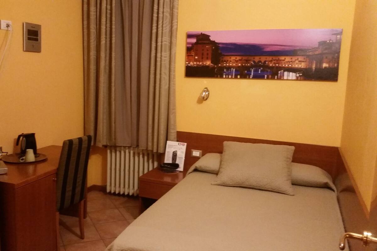 A hotel room with a bed, a desk, a chair, a radiator, a painting of a cityscape, and a telephone.