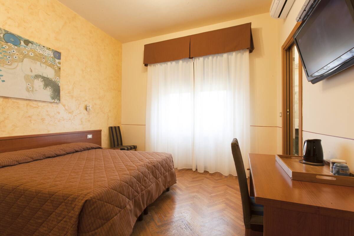 A hotel room with a large bed, a desk with a coffee maker, a chair, a television, and a window with curtains.
