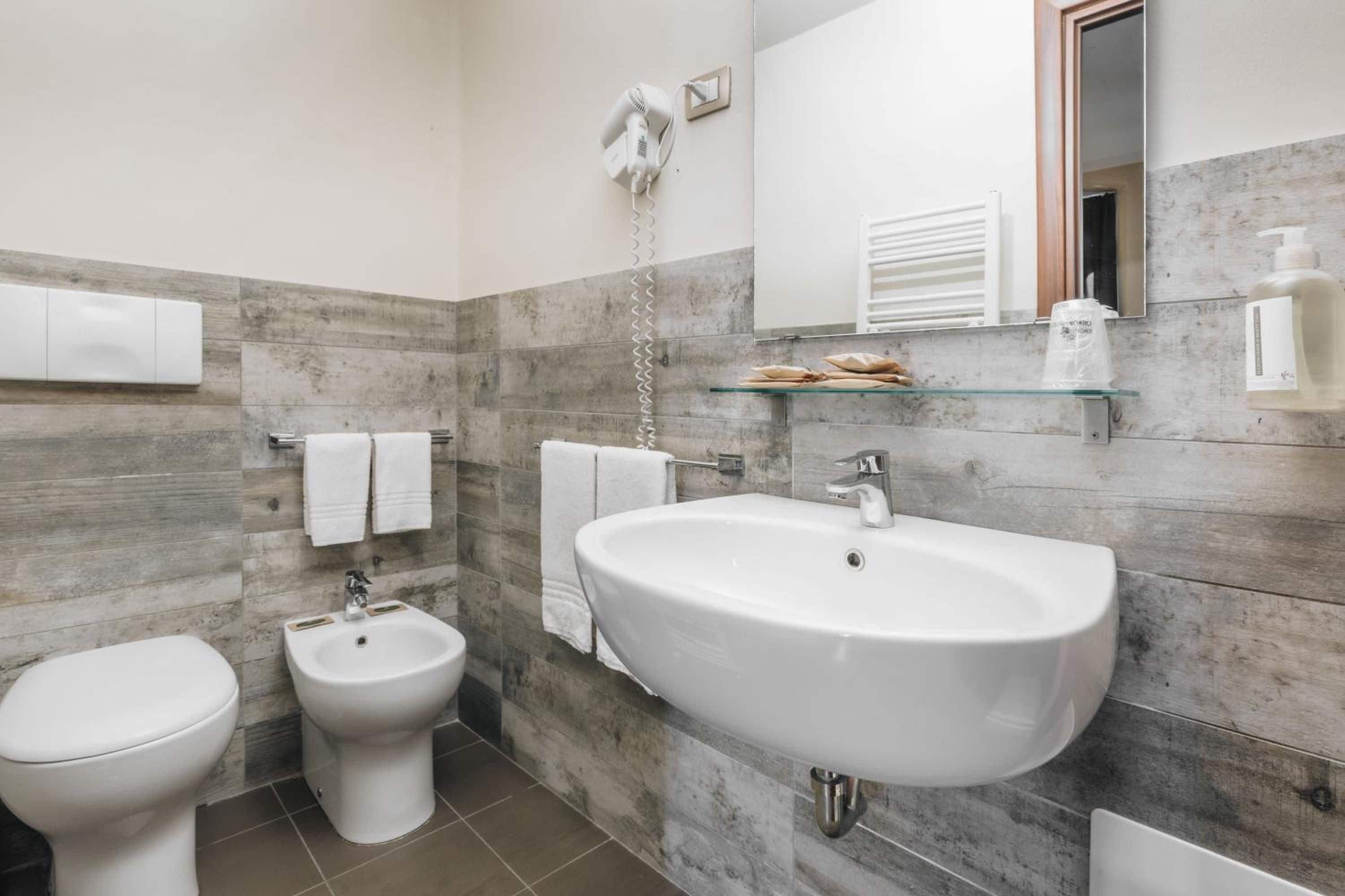 Ein Badezimmer mit einem weißen Waschbecken, einem Bidet und einer Toilette. Über dem Waschbecken befindet sich ein Spiegel und an der Wand ein Handtuchhalter.