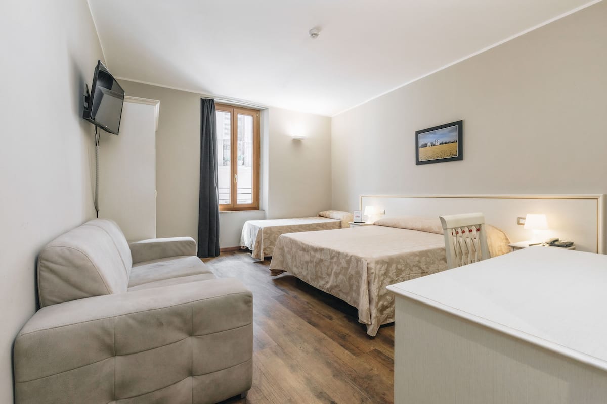 Habitación de hotel con sofá, dos camas individuales, escritorio, televisor montado en la pared y ventana con cortinas.