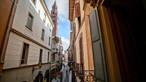 A narrow street with buildings on both sides, a prominent tower in the distance, and a metal railing on the right. - Hotel Astoria (Cremona)