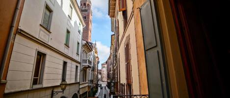 A narrow street with buildings on both sides, a prominent tower in the distance, and a metal railing on the right.