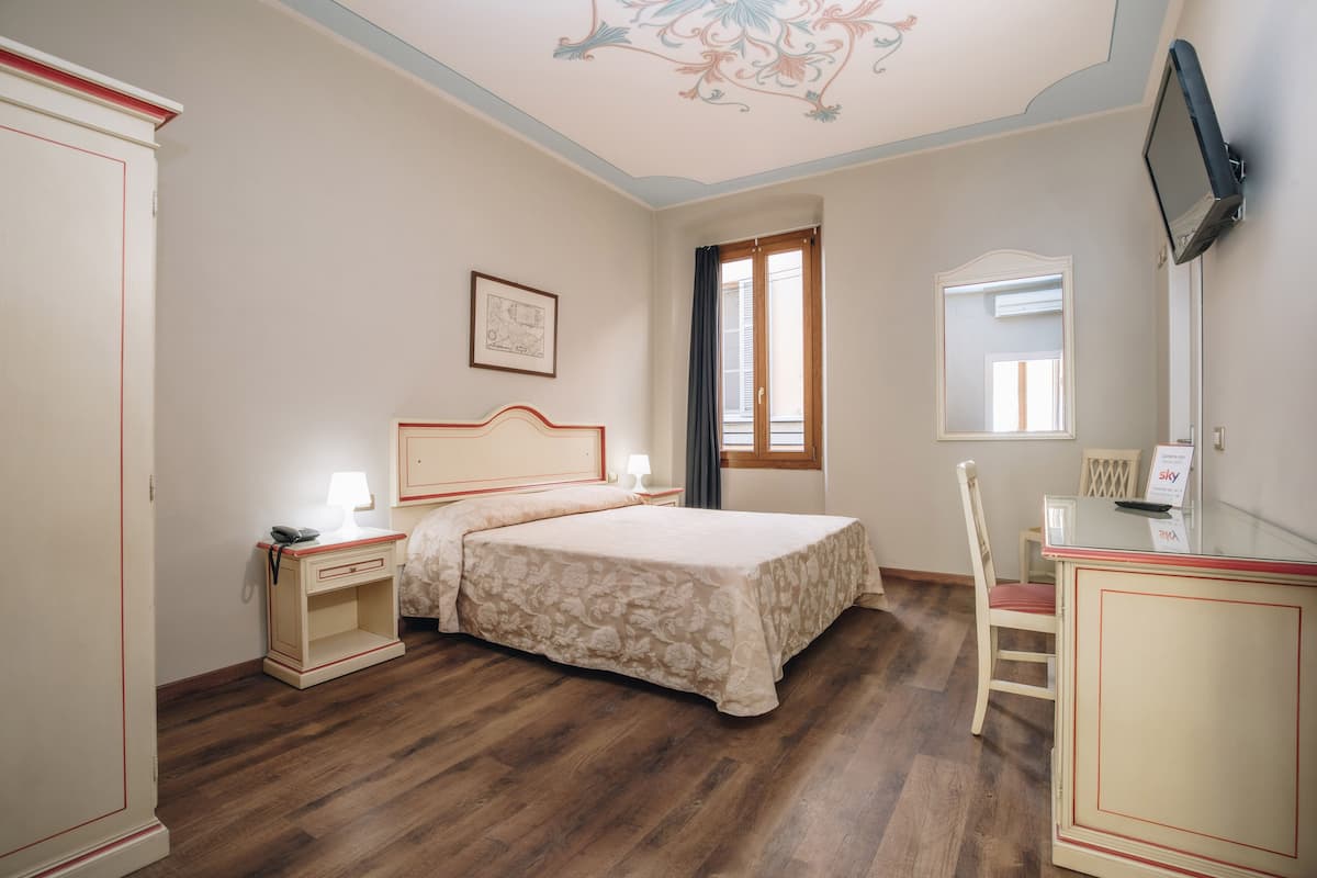 Un dormitorio con cama, mesitas de noche, escritorio, silla, ventana y televisor montado en la pared.