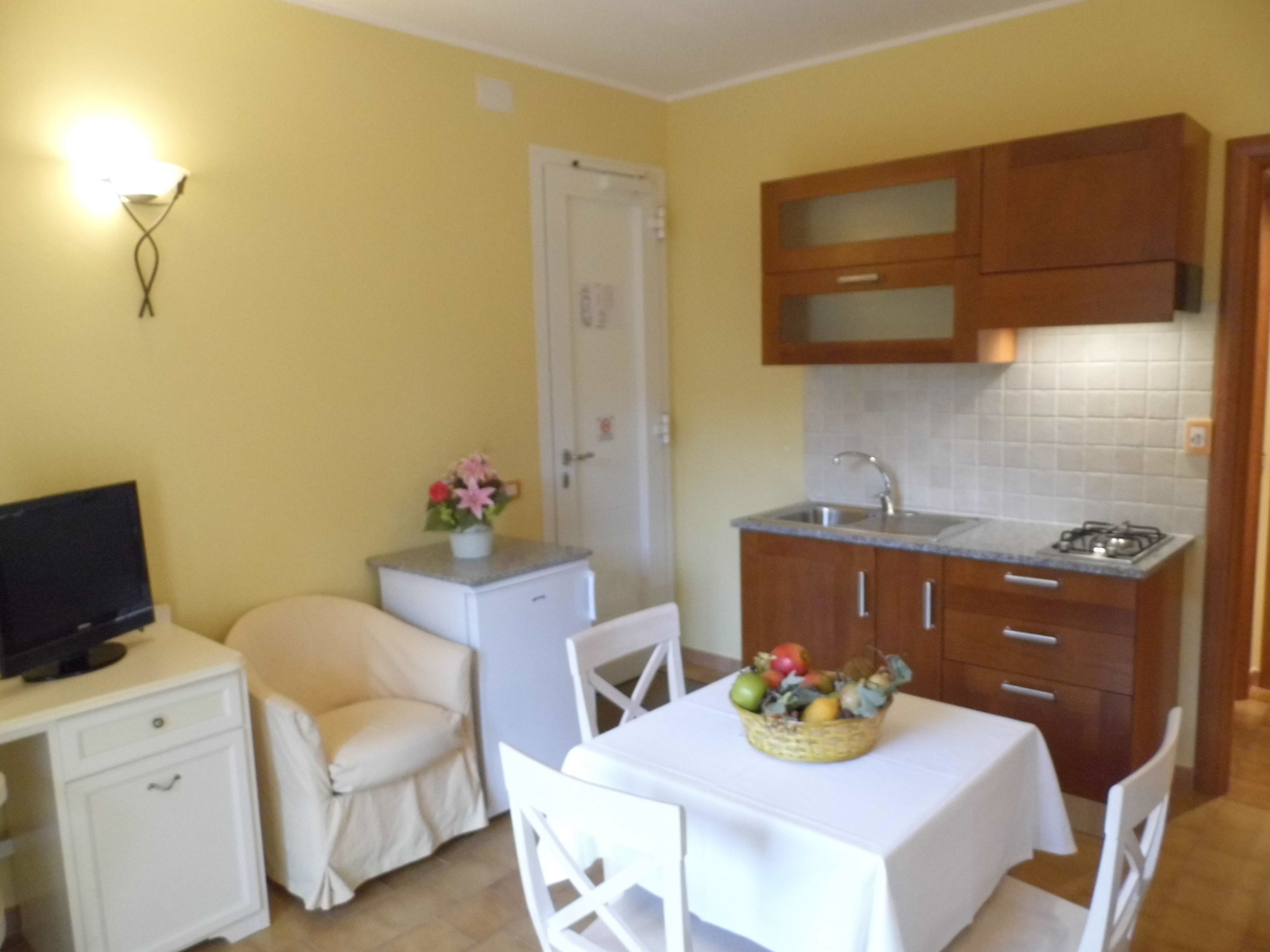 Une cuisine compacte comprenant un évier, une cuisinière et des placards, un petit réfrigérateur, un téléviseur posé sur une table de chevet, et un coin repas avec une table et des chaises.