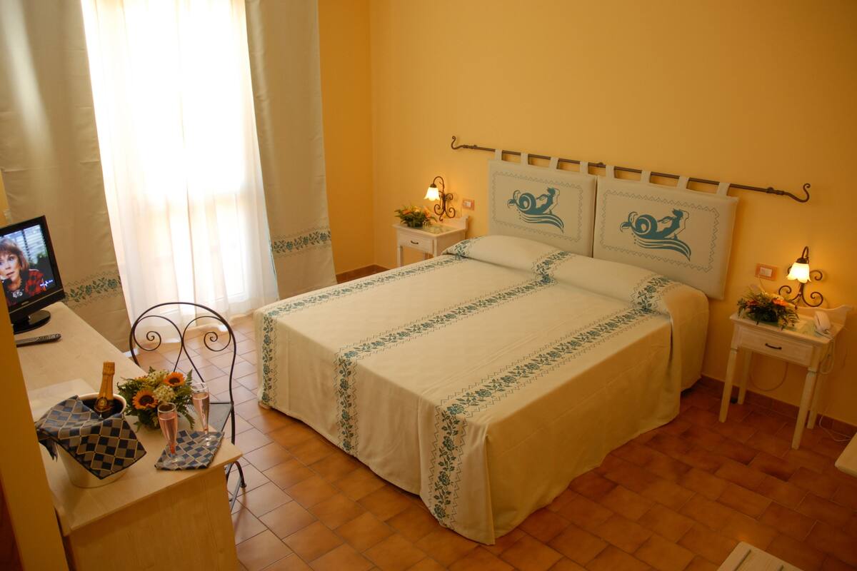 Un dormitorio ordenado con cama, mesitas de noche, un televisor y una pequeña mesa con una botella y vasos.