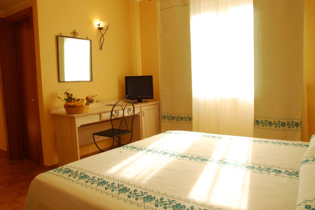 Una habitación de hotel con cama, escritorio con computadora, silla y ventana con cortinas.