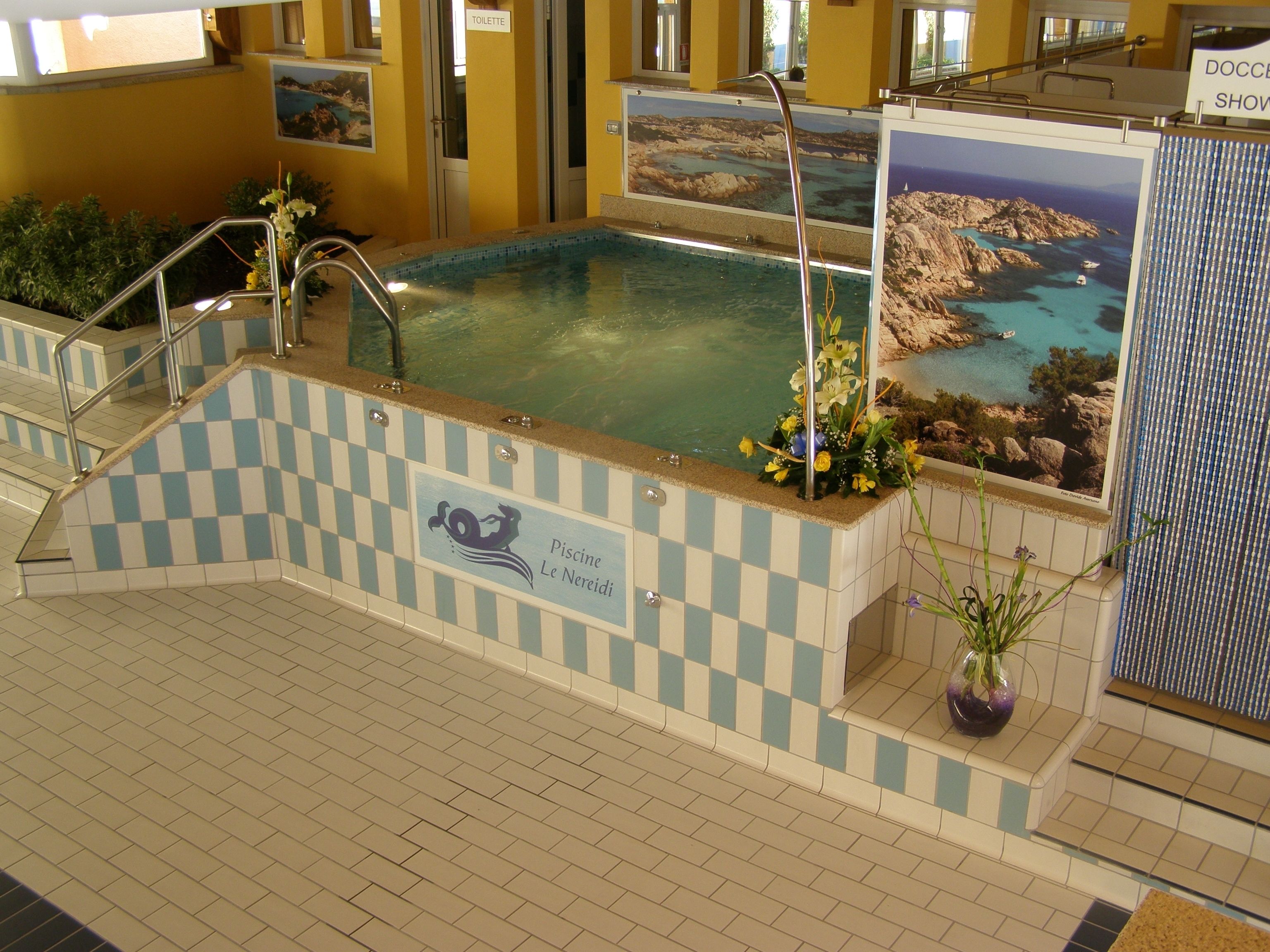 An indoor swimming pool with a checkered tile border, a sign indicating 'Piscine Le Nereidi', and a view of a coastal landscape through a glass wall.