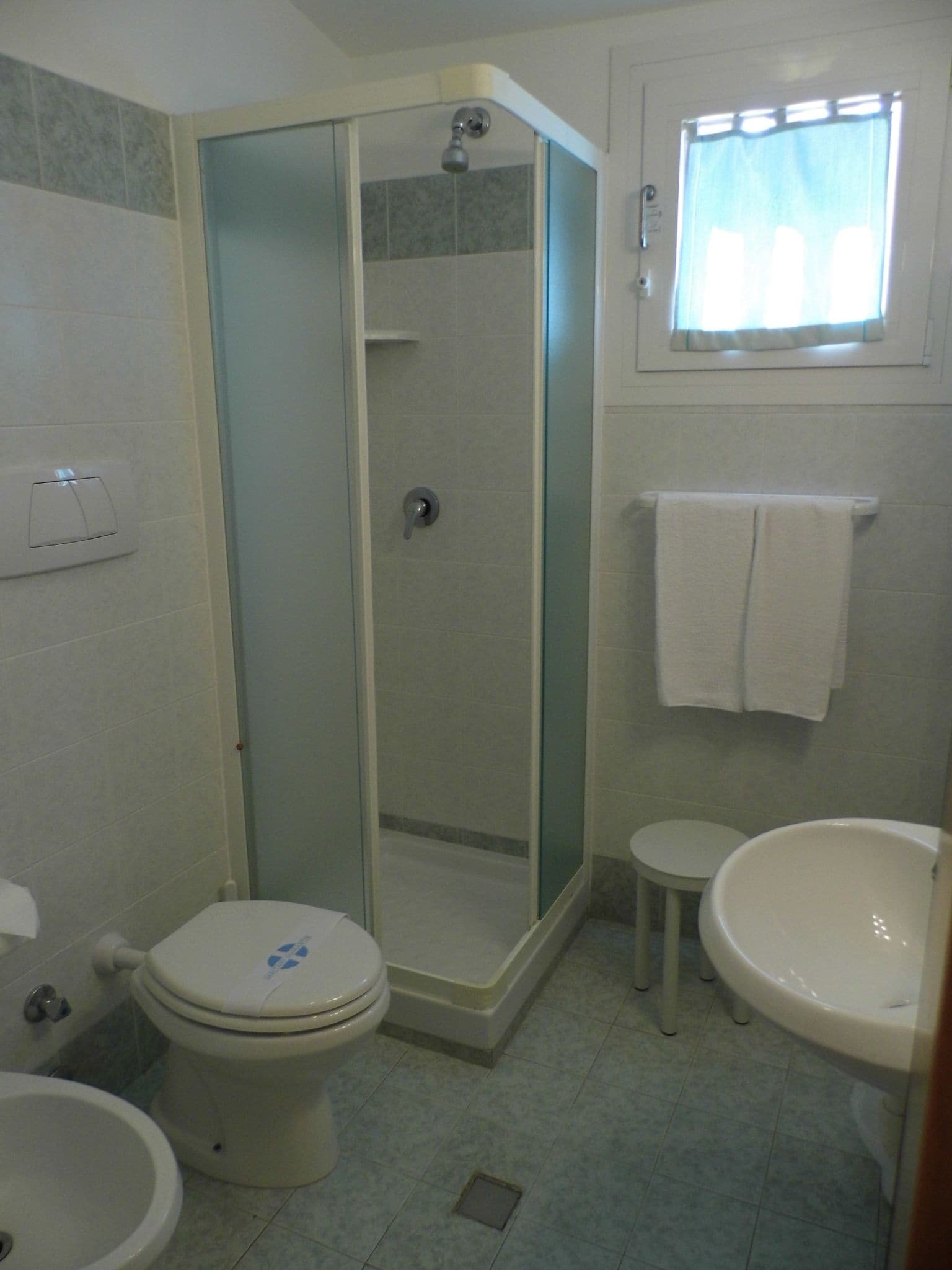 A bathroom with a glass-enclosed shower, a toilet, a sink, and a window with a curtain.