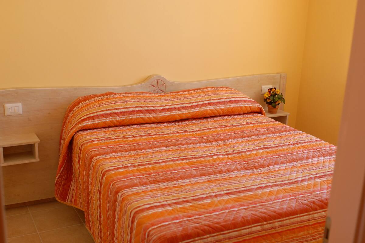 Una cama con colcha anaranjada y blanca a rayas, cabecera de madera y mesita de noche con un florero.