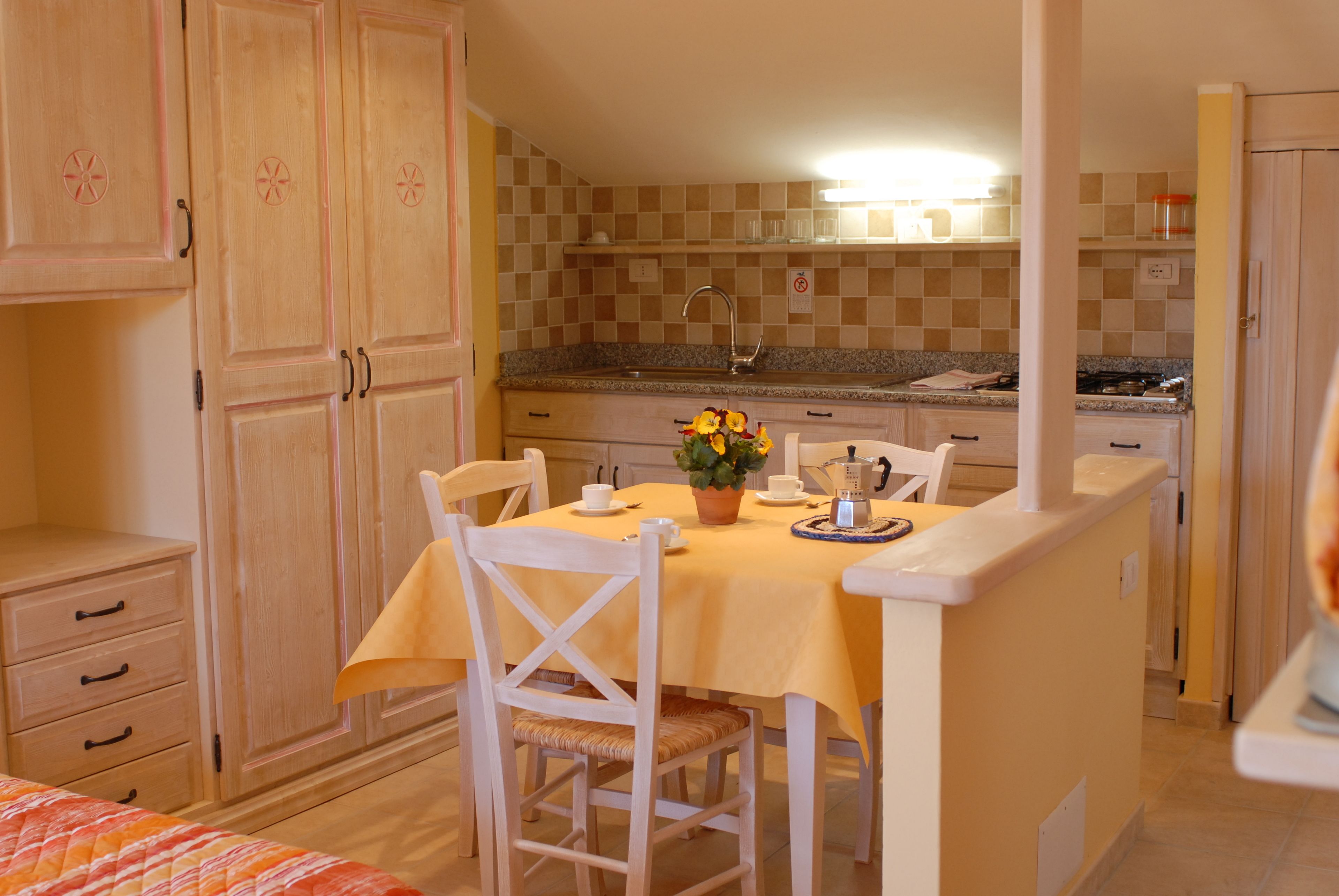 A compact kitchen with a dining area, featuring wooden cabinets, a checkered backsplash, and a small island with a yellow tablecloth.