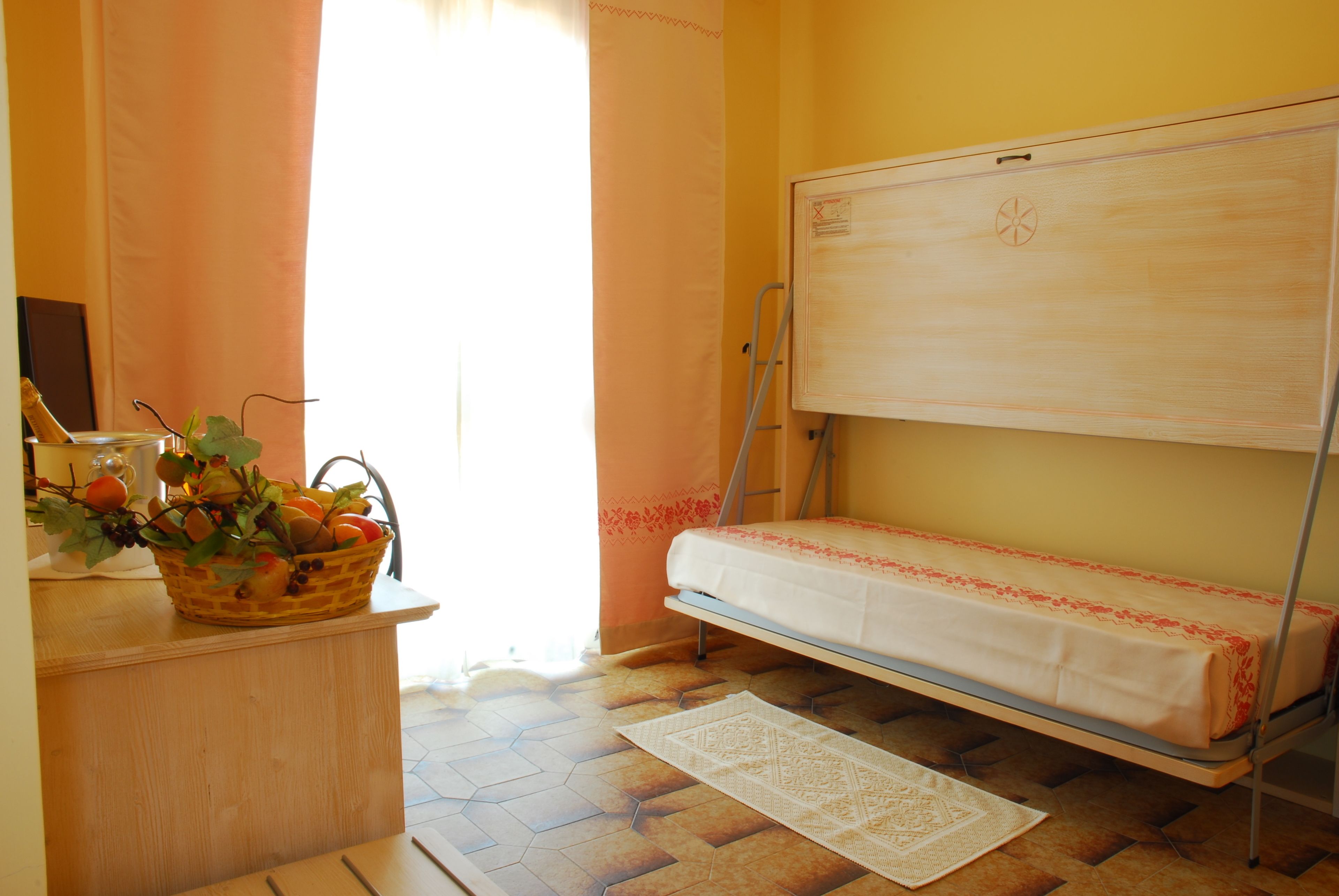 A room with a bed, a wooden desk, a basket of fruit, and a white rug.