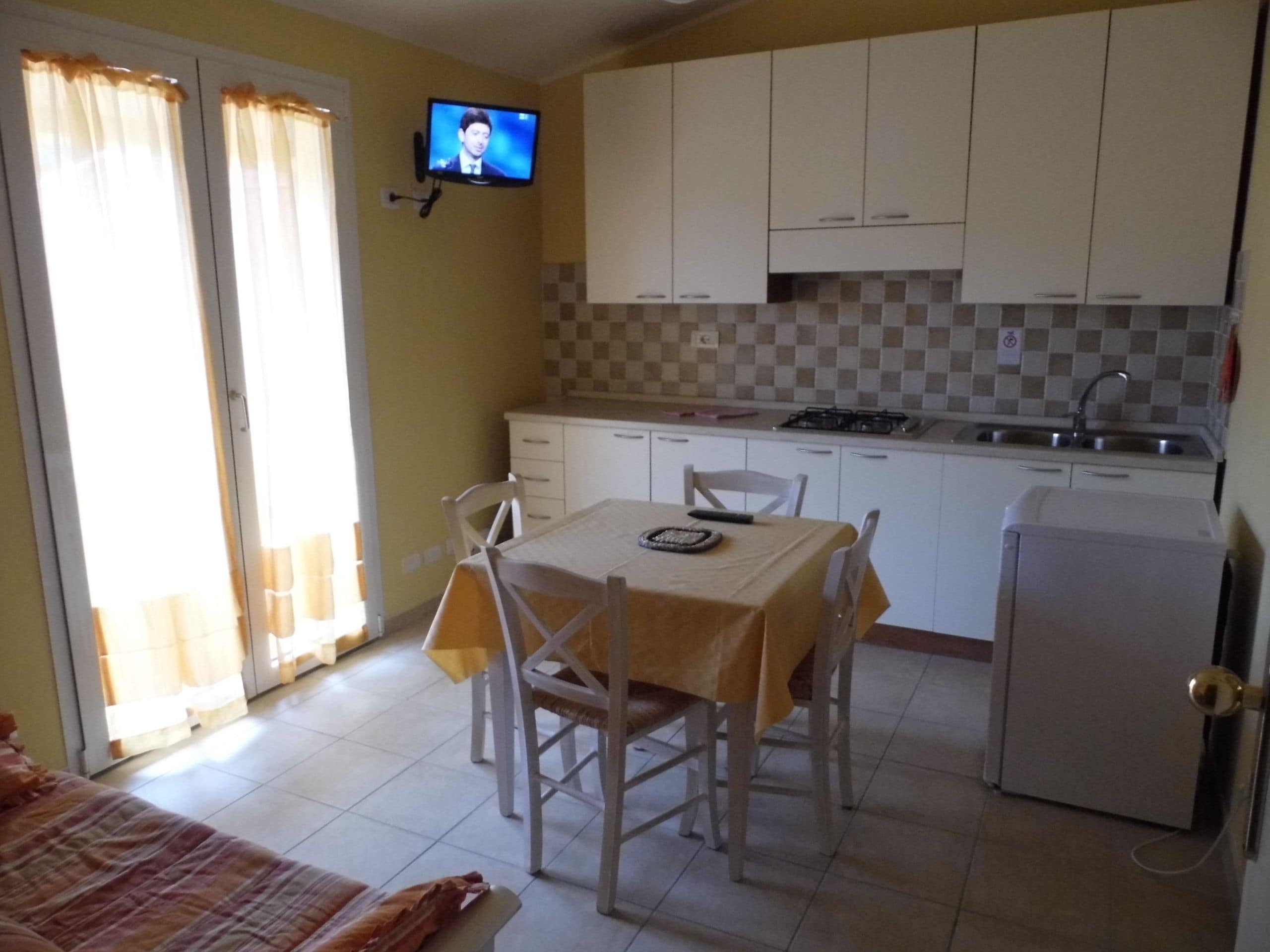 Une cuisine compacte avec des meubles blancs, une table à manger avec des chaises, un petit réfrigérateur et un téléviseur fixé au mur.
