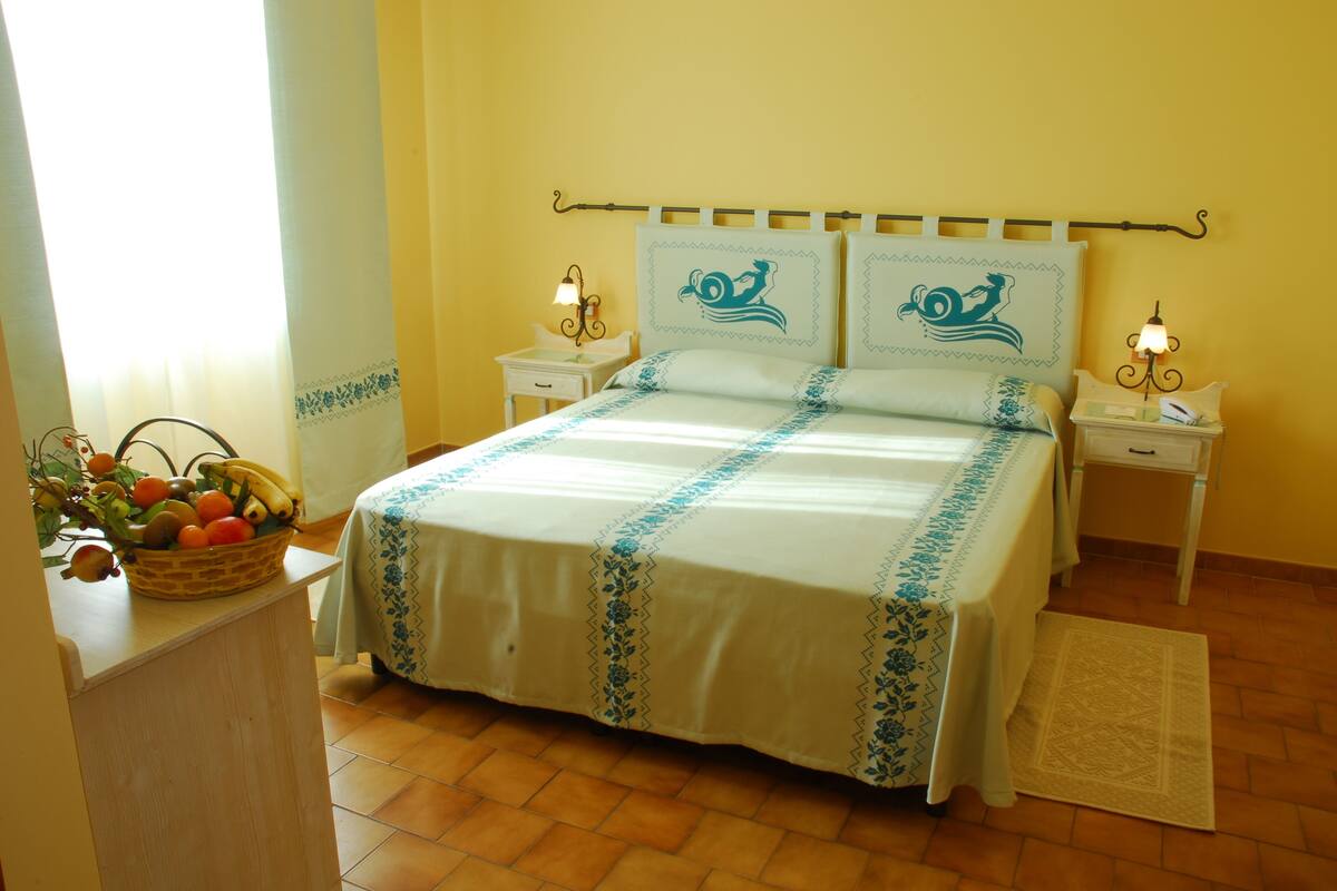 Un dormitorio ordenado con cama, dos mesitas de noche, una mesilla auxiliar, una cesta de frutas y una ventana con cortinas.