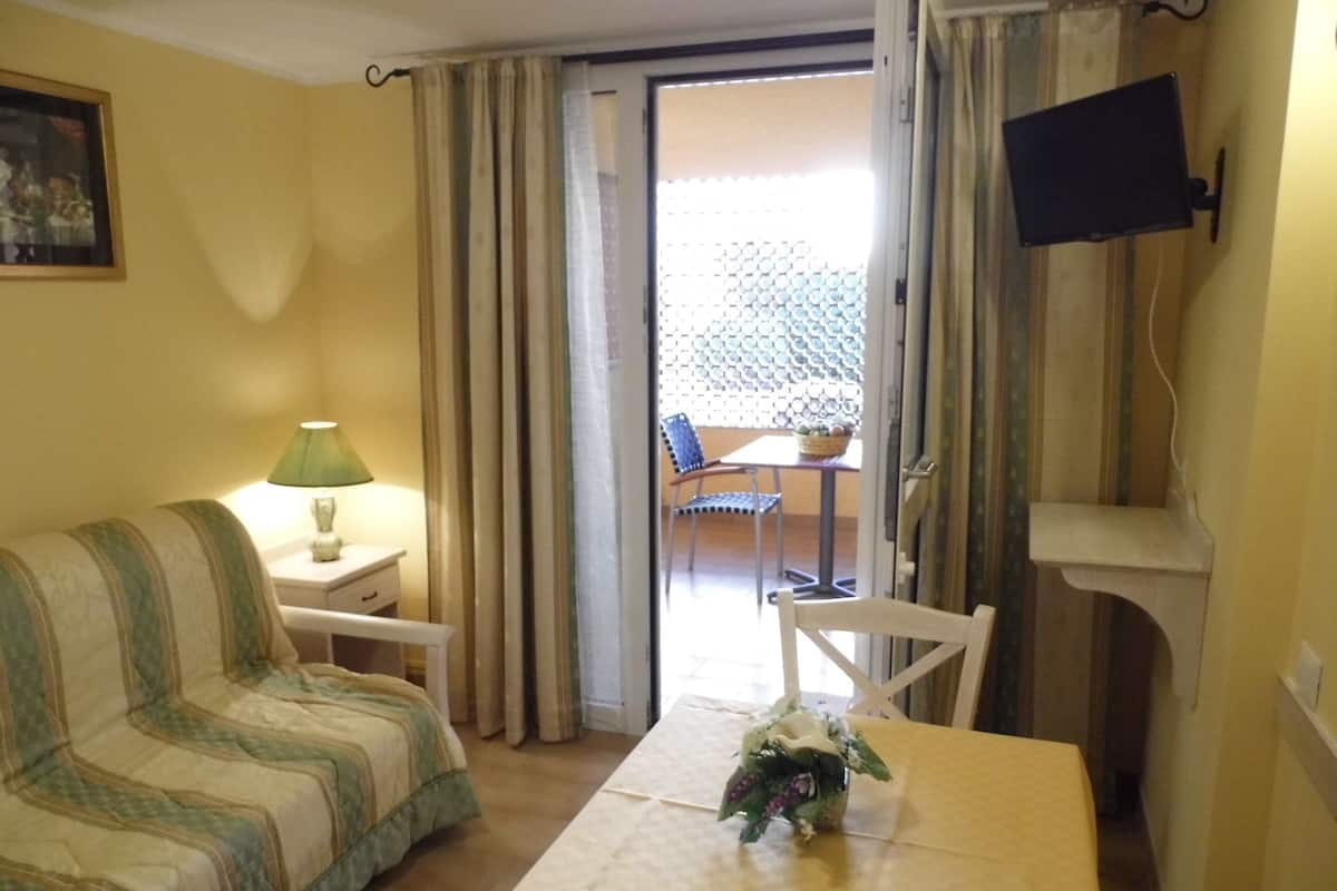 Una habitación con sofá, una mesita con un jarrón, una silla, un televisor montado en la pared y un balcón con una silla y una mesa.