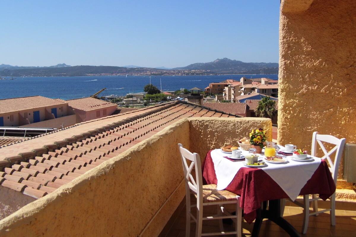 Un balcón con una mesa dispuesta para dos, con vista a un pueblo costero con edificios y un mar azul cristalino.