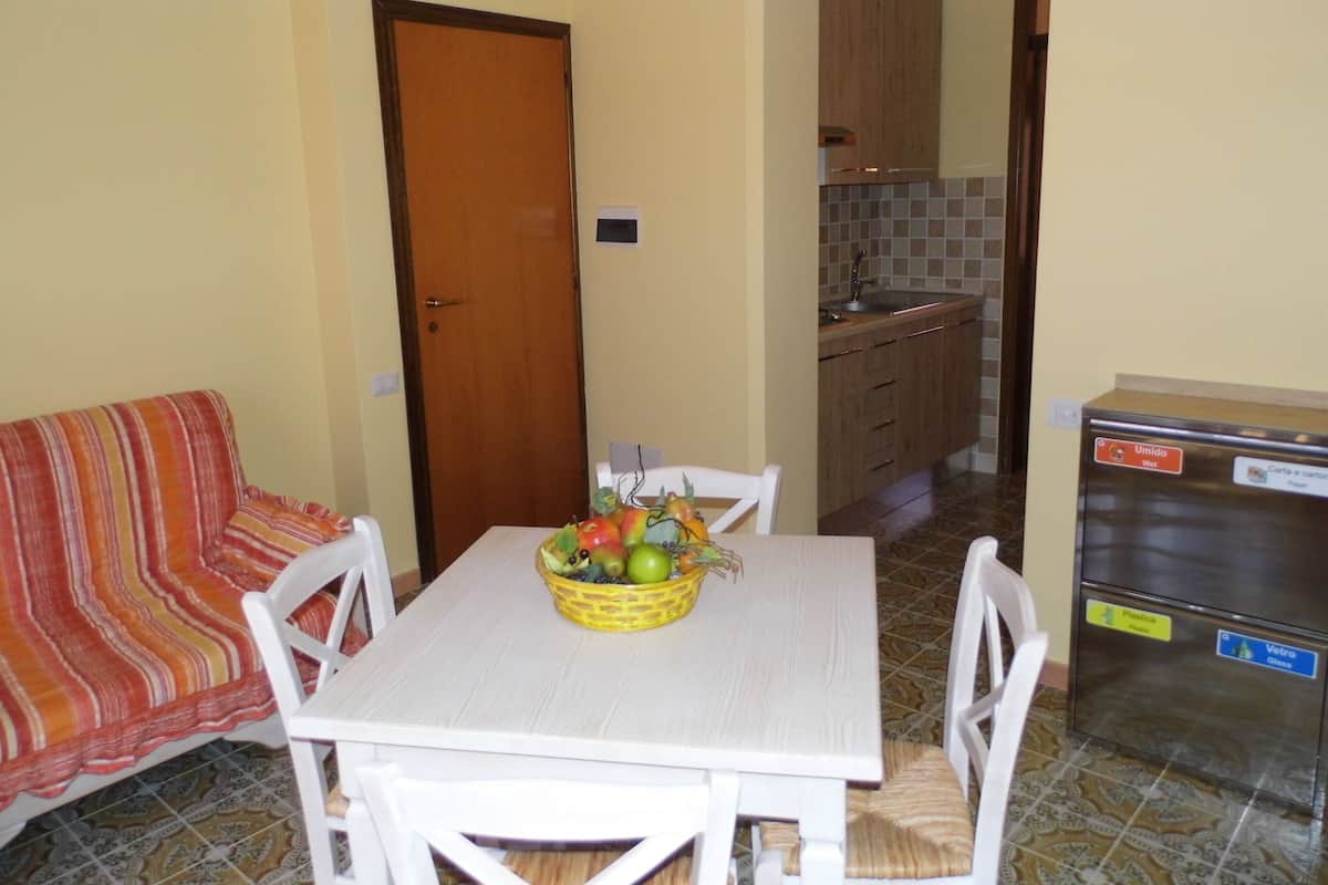 Un pequeño comedor con una mesa, sillas y una cesta de frutas. Se aprecia una cocina al fondo con armarios y un refrigerador.