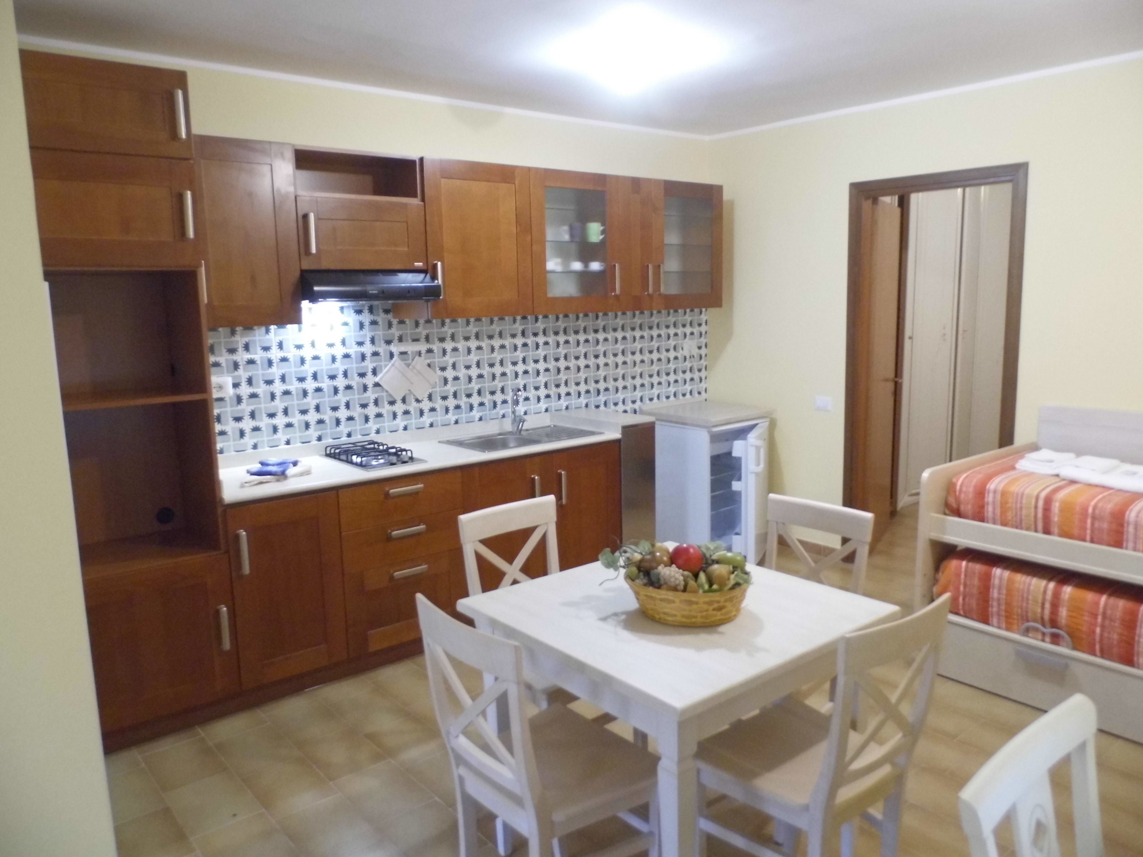A compact kitchen with wooden cabinets, a white countertop, and a tiled backsplash. There is a small refrigerator, a sink, and a dining table with chairs. A bed with a striped blanket is visible in the background.