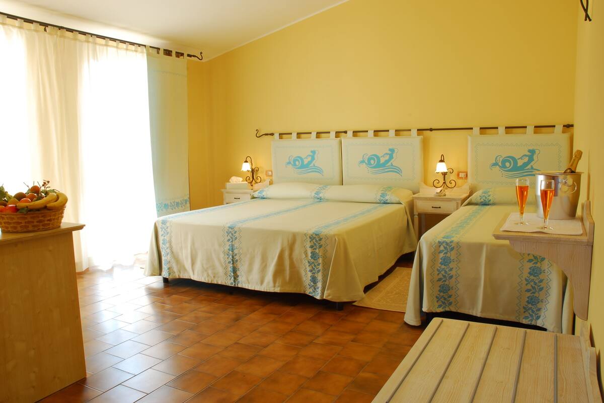 Habitación de hotel con dos camas, una mesita de noche de madera con una lámpara, una pequeña mesa con dos vasos de zumo de naranja y una cesta de fruta sobre un mueble de madera.
