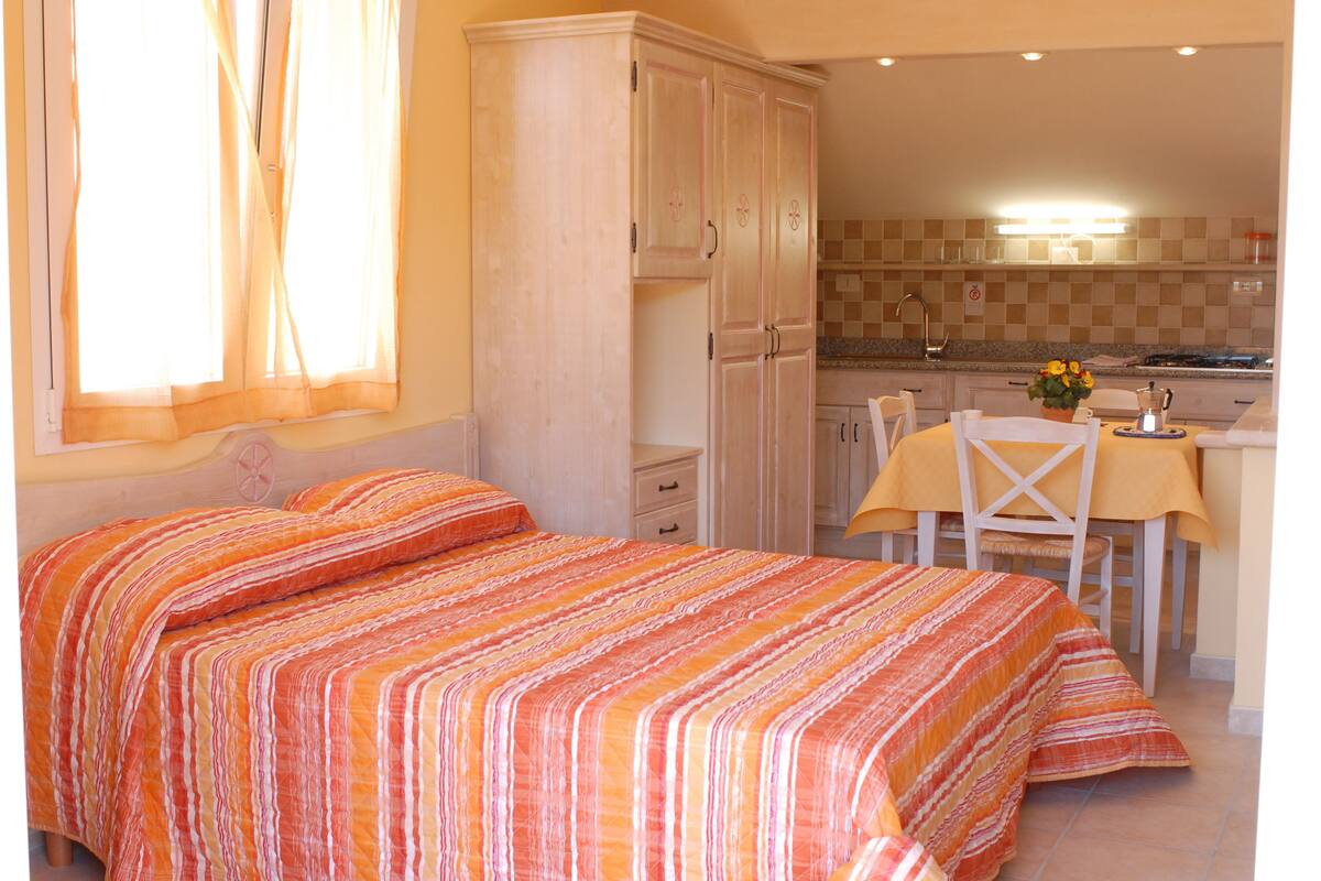 Un dormitorio con cama, armario, zona de comedor con mesa y sillas, y cocina al fondo.